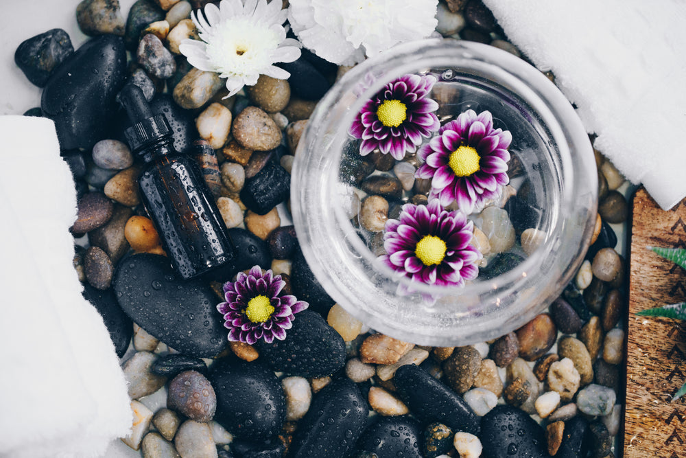 Glass bowl with purple flowers and essential oil bottle on smooth river stones — symbolizing Manna Oils’ natural skincare origins and mindful craftsmanship.