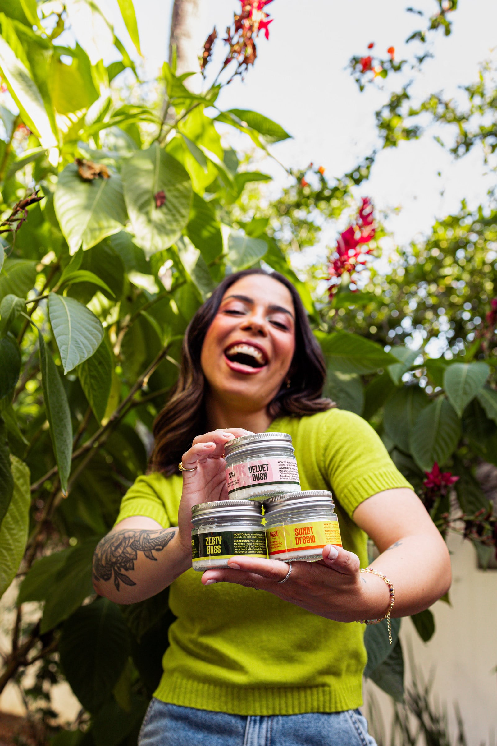 Miriam, founder of Manna Oils, laughing as she holds the three whipped body butters, Zesty Bliss, Velvet Dusk and Sunlit Dream, in a leafy garden