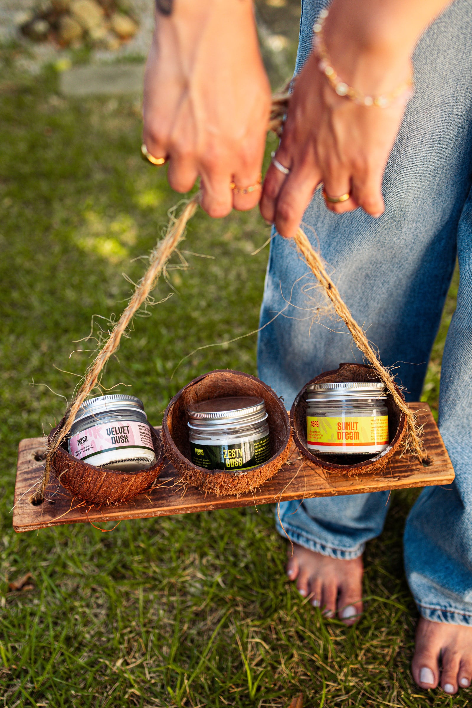 The three Manna body butters carried in coconut-shell holders on a wooden tray, over grass